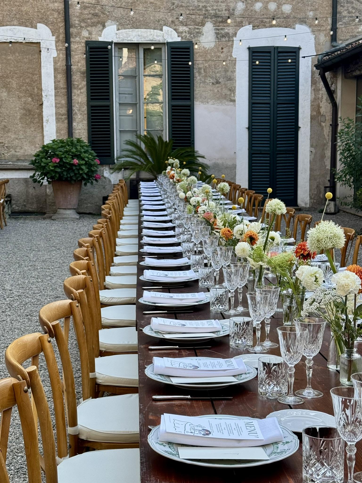 Lunga tavolata imbandita in un cortile storico, con luci calde e composizioni di dalie e ortensie